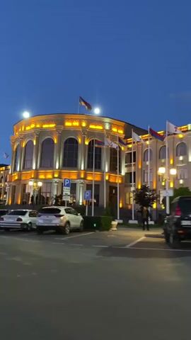 The video captures an exterior view of a grand building illuminated by warm, golden lights against a deep blue twilight sky. The building features classical architecture with arched windows and decorative molding, topped with flags, including the Armenian flag. Several cars are parked in front of the building, and a white minibus is seen driving by. The scene appears to be in a public space, possibly a government building or a cultural center. The time of day is dusk, as indicated by the dark sky with some remaining light on the horizon and the artificial lighting of the building. The weather appears clear. The overall impression is one of elegance and civic pride. No visible text is present in the video.