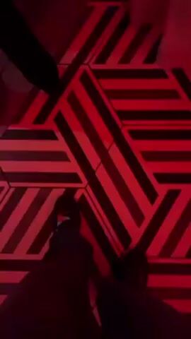 The video shows an energetic, dimly lit outdoor event, likely a nightclub or party. The floor is decorated with a striking geometric pattern of black and white stripes. People are milling around, some dancing, suggesting a lively atmosphere. The space is illuminated with red lighting, and in some shots, you can see modern buildings in the distance against the night sky. The video focuses on the energy and setting of the event, capturing movement and sounds.