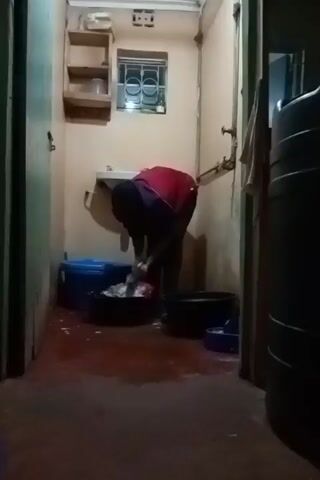 The video shows a person doing laundry in a small, dimly lit bathroom. The person, wearing a red shirt, is bent over a black tub filled with clothes, washing them in the sink. There are several other containers around, including a blue bin and another black tub. A water tank is visible on the right side of the frame. The floor is wet with a reddish tint. A small window provides some light. A child can be heard speaking in the background.