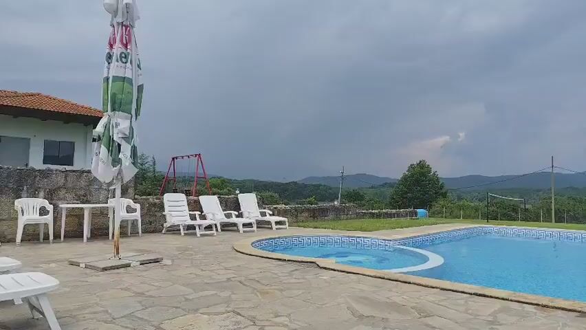 The video opens with a wide shot of an outdoor swimming pool area under a cloudy sky. The pool is blue and has a decorative design on the bottom. To the left of the pool, there are three white lounge chairs and an umbrella, with a table and chairs visible further right. In the background, there are rolling green hills and trees under the dark clouds. The camera slowly pans to the right, revealing more of the pool area and the surrounding landscape. As the camera pans, a table with four chairs and another umbrella come into view on the right side of the pool. The hills and trees in the distance remain visible, with the cloudy sky still overhead. The video ends with a view of the pool and the landscape as the camera completes its pan.