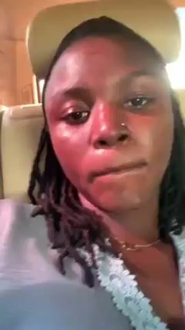 The video shows a woman with dark skin and black dreadlocks. She is wearing a light-colored shirt with lace on the collar, a gold necklace, and a light-colored baseball cap. She has a nose ring. The woman looks directly at the camera. She raises one hand and extends two fingers into a V shape, or peace sign. The background appears to be the interior of a car.  There is a headrest visible behind her. There is an out-of-focus voice speaking in a foreign language in the background.