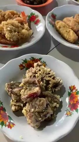 The video shows a table arrangement of several small plates with food. The plates are mostly white with colorful floral patterns. There are plates of fried food including what appears to be eggplant, shrimp, and some type of fried logs. There are also some saucy items in orange and green bowls, and a plate with mixed greens. The table top is light grey and smooth.