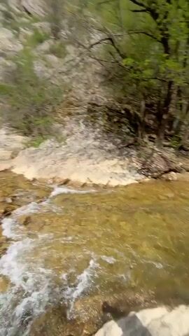 A young person in a black jacket and baseball cap is filming themselves with a phone in a rocky outdoor environment, holding a water bottle. Behind them, a waterfall flows into a clear pool of water. Several people are in the background, some sitting by the water and others taking pictures. The background features rocky cliffs and trees. The sky is partly cloudy. The person filming says “So beautiful”.