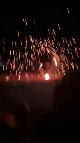 A person is spinning a flaming object in a dark outdoor setting. Numerous sparks are flying outwards from the spinning object, creating a circular spray of light. The ground appears to be dirt and sparse vegetation. The sky is black and clear. A person's shoulder and head are visible in the lower right corner of the frame, suggesting the video is being recorded from a close distance by an observer. The overall impression is of a fire performance or display.