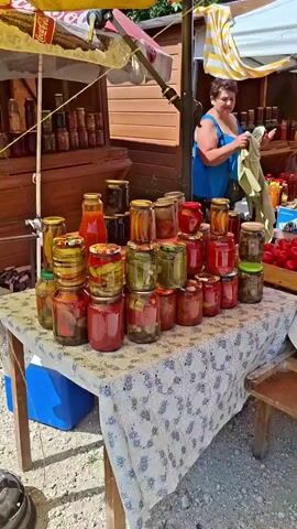 A vendor stands behind a market stall, holding up a light-colored garment. In front of the stall, a table is covered with a tablecloth adorned with a floral pattern. Displayed on the table are numerous jars of preserved food, including what appears to be pickles and tomatoes. To the right of the jars, several wooden crates are filled with fresh produce. One crate contains a large quantity of red tomatoes, another has a mix of dark-colored tomatoes and orange fruits, and a third crate holds beets with their green tops. A white bucket filled with green peppers sits on the edge of the table. Further back, more jars of preserved goods and bundles of greens are visible. The market appears to be set up outdoors, with wooden structures and an awning providing shade. Another person can be seen in the background behind the main stall.