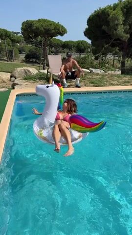 A person is floating on an inflatable unicorn in a swimming pool. Another person is standing at the edge of the pool, holding a beach ball and appears to be about to throw it. The sun is shining and the environment appears to be outdoors with trees and rocks in the background.