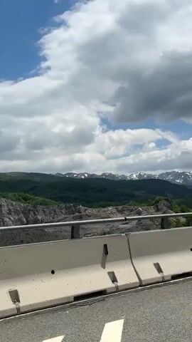 The video shows a view of mountains from a roadside. The camera is positioned on a road, with a concrete barrier visible in the foreground. Beyond the barrier, there is a rocky, barren area, followed by a dense green forest. In the distance, snow-capped mountains are visible against a partly cloudy sky. A tall, gray light pole is visible on the right side of the frame, occasionally obstructing the view of the landscape. The camera appears to be moving, as the light pole shifts position relative to the view.