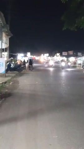 A nighttime street scene shows a paved road with a few motorcycles and a few people. There are buildings on the left side of the road, and stalls or shops are visible on the right side of the road. The sky is dark, and the scene is lit by artificial lights from the buildings and vehicles.