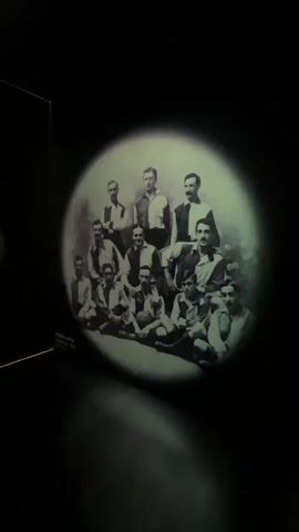 The video shows a black and white photograph projected onto a dark surface. The photograph depicts a group of approximately 13 men, presumably a sports team, posing for a formal portrait. The men are arranged in two rows: a back row of five standing men and a front row of eight seated or kneeling men. They are all wearing similar uniforms with distinct collars and stripes on their sleeves, suggesting a team sport. The lighting in the video focuses on the projected image, creating a spotlight effect and leaving the surrounding environment dark. The image is circular in shape, further emphasizing the spotlight effect. The video is short, lasting only a few seconds, and contains no discernible audio. The overall impression is that of a historical artifact or exhibit being presented.