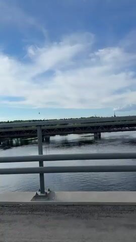 The video shows a wide shot of a river with two bridges spanning it under a bright blue sky with scattered white clouds. The camera appears to be on a road bridge, with a grey metal railing and a concrete surface with a drain visible in the foreground. Looking across the river, a dark grey metal truss bridge, likely a railway bridge, is visible in the middle ground, with its structure reflected in the water. A faint sound of a train passing on this further bridge can be heard around the 0:02 mark. Green trees and foliage line the distant riverbanks. A small white object, possibly a bird or kite, is seen briefly in the sky above the distant bridge around 0:01. The scene is brightly lit, suggesting daytime. The location is Imatra, Finland.