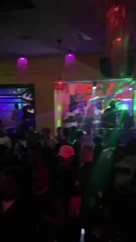 The video shows a crowded nightclub. There are numerous people dancing and talking near a DJ booth. A DJ is actively performing, and colorful lighting illuminates the scene. Decorative hanging lights and tropical plants are visible, adding to the ambiance. The camera is hand-held and moves slightly.