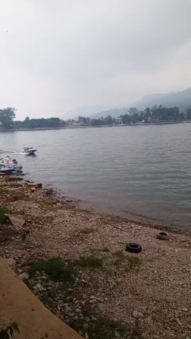 A rocky shore with two tires visible is in the foreground. A large body of water stretches into the distance, with a boat moving on the water in the middle ground. Trees and buildings are visible on the far bank, with a misty mountain range in the background under a cloudy sky.
