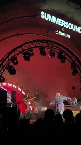 A live band performs on an outdoor stage at a festival. The stage is illuminated with red lights, and a large banner above the band reads "SUMMERSOUND" with a logo. The band consists of at least five members, including guitarists, a singer, and a keyboardist. The singer, a woman with blonde hair, is center stage, singing and dancing. A crowd of spectators is visible in the foreground, silhouetted against the stage lights. The overall atmosphere appears to be energetic and festive.