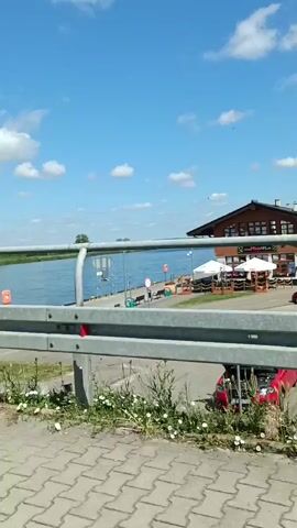 The video opens with a view of a blue sky with scattered white clouds, overlooking a body of water. A grey metal railing is in the foreground, with some green vegetation growing beneath it. Beyond the railing, there is a paved walkway or road next to the water, and a building with a brown facade and a red roof. Several white umbrellas are set up outside the building.

The camera then pans to the right, showing a coral-colored metal fence along the walkway. Two people are walking on the walkway, one in a dark shirt and the other in a brightly colored shirt and shorts.

The camera continues to pan, revealing more of the walkway and the water. Another building, this one with a white facade, is visible in the distance.

A large, light green metal pillar comes into view in the center of the frame. The camera continues to pan to the right, showing more of the railing and the water.

Finally, the video shows a person riding a bicycle across the scene from right to left. The cyclist is wearing a helmet and a patterned jacket. The bicycle has a blue bag attached to the handlebars. The water and the sky remain visible in the background. The video ends with a close-up of the bicycle's front wheel.