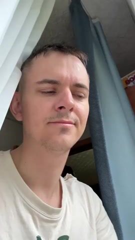 The video shows a man standing in front of a window with curtains. He is wearing a light-colored t-shirt and has short, dark hair. He holds up a flip phone to his ear and says "Allo" twice. A poster with the word "BOOM" is visible on the wall behind him.