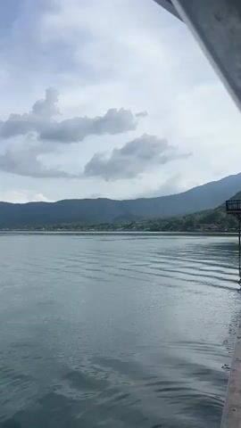 A static medium outdoor shot shows a large body of water extending across the bottom and middle of the frame, with green mountains in the distance and a cloudy sky above. The water has ripples, and the mountains are covered in trees. On the far left, a red buoy is visible. The camera slightly pans to the right from 0:03 to 0:04, then back to the left from 0:04 to 0:05.