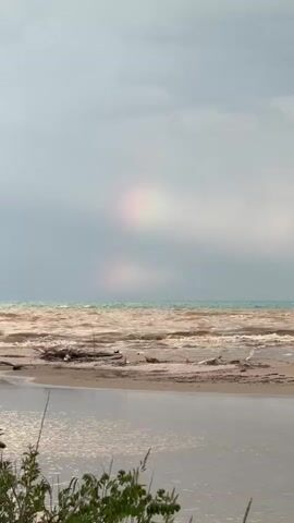 The video shows a landscape of a beach on an overcast day. The water is a murky brown color with visible waves breaking on the shore. Debris, including driftwood, is scattered along the beach. In the foreground, there's a smaller body of water connected to the larger body. Green vegetation is visible along the edges of the frame. In the distance, a faint rainbow is visible through the heavy clouds. The camera gradually zooms out to reveal more of the sandy ground and vegetation leading up to the beach. The overall mood is somewhat bleak due to the stormy sky and murky water.