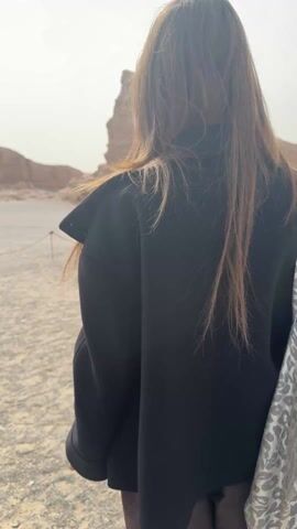 A young woman with long brown hair, wearing a black jacket, dark trousers, and dark shoes, and carrying a white and gray patterned tote bag, is seen walking and posing in a desert landscape. The scene features a vast, gravelly ground with distinctive large rock formations (likely yardangs) under a bright, slightly overcast sky. Simple rope barriers delineate the walking path.

At 0:00, she walks away from the camera, revealing the impressive rock formations in the background. Around 0:01, she turns to her left, observing the scenery. By 0:02, she faces the camera, smiles, and spreads her arms wide with her hands raised, striking a pose. She then begins to walk towards the camera, maintaining a happy expression. The sunlight casts clear shadows, indicating it is daytime. The overall mood is one of enjoyment and exploration in a natural, rugged environment. The location cues are consistent with the Yardang National Geological Park in Dunhuang City, China.