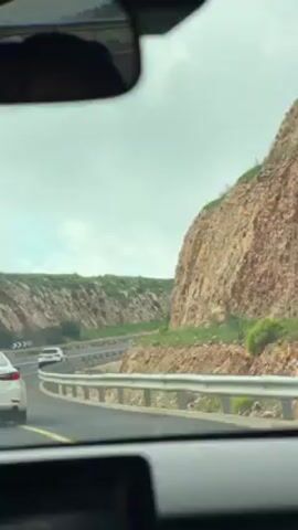 The video shows the view from inside a car driving on a highway. The road curves slightly to the left, and there are other cars on the road in the same direction. A metal guardrail separates the road from the side of a rocky hill. The sky is overcast. The rear view mirror is visible at the top of the frame.