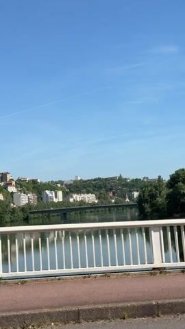 The video captures a daytime scene from a bridge overlooking a river, with buildings visible on the opposite bank. The sky is clear and blue. A person on a red bike and another on a scooter pass by. The bridge has white railings and a red walkway.