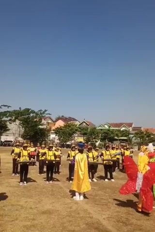 A group of young people in matching yellow vests and black pants are standing in formation on a grassy field under a clear blue sky. They are holding instruments, possibly drums or cymbals, and appear to be part of a marching band. A person in a yellow cape and a blue hat stands in front of them, facing the camera. In the background, there are houses and trees. The overall scene suggests an outdoor performance or rehearsal.