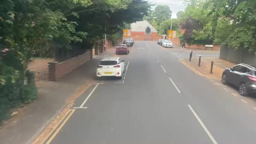 A white car is parked on the left side of a paved road. The car is angled towards the street, with its front facing away from the camera. There are trees and bushes on the left side of the road, as well as a brick wall. On the right side of the road, there are more trees and bushes, a few parked cars, and a building in the background. The sky is overcast. There is a speed limit sign that reads 20 zone with a symbol of a child walking.