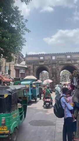 A street scene unfolds with several auto-rickshaws visible, one a vibrant green in the foreground. Pedestrians are present, including a man carrying a young child on his shoulders, his back to the camera. In the background, a large arched gateway, constructed from stone, stands prominently. The sky above is partly cloudy. The overall impression is one of a bustling market or thoroughfare in a location with historical architecture.