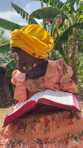 A young girl, likely of African descent, is seated outdoors holding a large book with a bright red spine and edges. She is wearing a peach-colored dress with floral print and a yellow head wrap. Her face is adorned with white markings, including a heart symbol on her forehead. She is looking down at the book, then glances to her right, and then down again. The background features lush greenery, including banana trees, suggesting a tropical or rural setting. A building or structure is faintly visible behind the foliage.