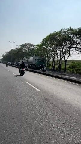 A motorcycle rider films the road ahead while traveling on a sunny day. The rider is wearing a black helmet and a dark jacket. Several other motorcycles and cars are visible on the road. A large tanker truck and a white truck are stopped on the side of the road, appearing to be part of a traffic jam. Trees line the side of the road.