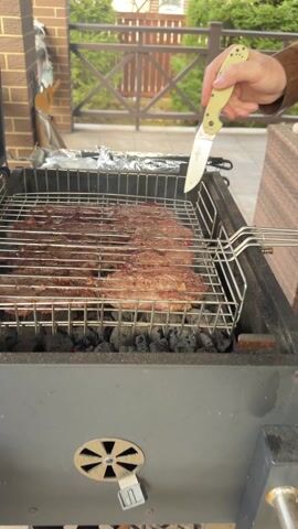 A video shows meat cooking in a grill over charcoal. A person's hand is visible holding a knife, but they are not interacting directly with the food or grill. The grill is positioned on a patio with a brick building and a wooden fence visible in the background. The meat looks well-seasoned and is contained within a metal grilling basket.