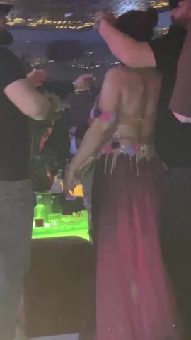 The video depicts a person dancing on a table in what appears to be a nightclub or party setting. The dancer is wearing a semi-transparent outfit adorned with floral decorations and a long, pinkish train. Several people surround the dancer, some of whom are taking photos or videos with their phones. A table in the foreground features bottles and glasses, illuminated by colorful, likely LED lighting. The overall atmosphere seems lively and festive. The background is filled with other patrons and dim lighting, typical of such venues.