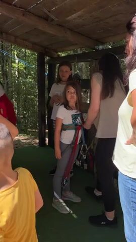 A full shot shows four individuals standing within a wooden structure with an open side looking out into a forest. The individual on the far right wears blue jeans and a white short-sleeved top, partially obscured by the individual to her left. This second individual wears dark pants and a beige short-sleeved top and leans over to assist a young girl in the center. The young girl wears a white t-shirt, grey leggings, and a harness, with long brown hair. Another young girl, with long brown hair and wearing a white t-shirt and plaid pants, stands behind the harnessed girl. A young boy in a red short-sleeved shirt is partially visible on the far left. The ground is covered with green matting.