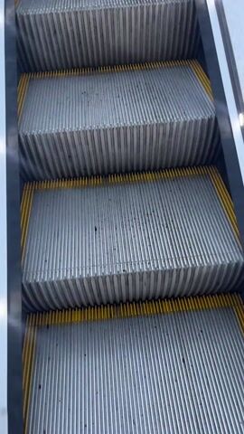 The camera is pointing upward at a slowly moving escalator with gray metal steps and yellow sides, as it ascends. The escalator has glass side panels with silver-colored rails and lights running along the rails. The background shows an overcast sky and other buildings.
