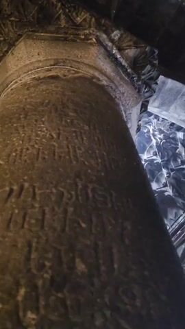 A brief, shaky video pans up from the base of a stone pillar, showing carvings on its surface, to reveal a highly ornate, vaulted ceiling with a bright light shining from an opening at the apex. The pillar appears to be old and weathered, with inscriptions etched into its surface. The ceiling is a complex geometric design, possibly made of stone or stucco, with deep shadows and dramatic highlights due to the intense light from above. The overall impression is one of an ancient, possibly religious or historical structure, with a sense of awe and wonder inspired by the architecture and the light.