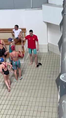 The video shows a group of swimmers and two coaches at an indoor pool. The swimmers, dressed in swimsuits and caps, are standing near the edge of the pool. One coach, wearing a red shirt and blue shorts, is barefoot and talking to them, gesturing with his hands. The other coach is sitting on a wooden bench, wearing a white shirt and brown shorts, and looking at a paper in his hand. Several pairs of shoes and a red towel are lying on the bench. The floor is covered with white and gray square tiles. The walls are white, with a staircase-like structure in the background. The camera is positioned at an elevated angle, looking down at the scene.