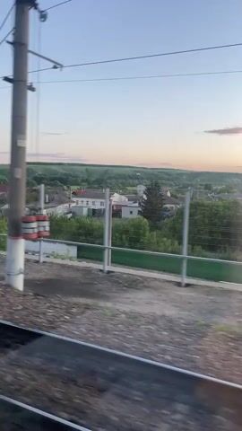 The video captures a view from a moving train, looking out over a landscape at sunset. In the immediate foreground, blurred train tracks with gravel ballast are visible, indicating the motion of the viewing platform. A grey utility pole with red and white banding, and associated overhead wires, quickly passes on the left side, appearing blurred due to speed.

Beyond the tracks, a low chain-link fence runs parallel to the railway. In the midground, a residential area spreads across gently rolling hills, featuring numerous light-colored houses with varying roof styles, interspersed with abundant green trees and foliage. As the train continues to move, a curved road with several cars driving on it comes into view, stretching across the middle distance. Streetlights or similar structures line the road.

The sky above transitions from a pale blue at the top to soft hues of orange and yellow towards the horizon, characteristic of a sunset. A few small, wispy pink clouds are scattered across the sky. In the far distance on the right, a distinctive white building with domes, likely an Orthodox church, is visible against the fading light.

The overall scene depicts a peaceful, late afternoon or early evening setting in a rural or suburban area. The location is Zaluzhnoye, Russia. No visible text is present.