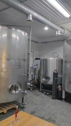 The video pans across a room with industrial stainless steel tanks and brewing equipment. The room appears to be a brewery or similar production facility. The tanks are large and cylindrical with various valves and pipes attached. There are also some smaller tanks, gas cylinders, and bottles visible. The lighting is fluorescent and the ceiling is corrugated metal. The floor is concrete.