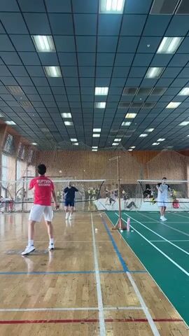 This is a vertical, first-person perspective video of a badminton match in an indoor gymnasium. The camera is positioned on the court, looking towards the net. The court has a wooden floor with blue and white markings. Several people are visible playing badminton. In the foreground on the left, a person in a red t-shirt and white shorts is facing away from the camera, holding a badminton racket. On the right side of the net, another person in a white t-shirt and shorts is also facing the net. In the middle ground, several other players and spectators are visible. The gymnasium has a tiled ceiling with fluorescent lighting. The game appears to be in progress, with players actively engaged in hitting the shuttlecock.