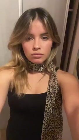 A young woman is taking a selfie video in front of a closet or door. She is wearing a black top and has a leopard print scarf around her neck. Her hair is long, with blonde highlights, and she has bangs. She touches her hair in the video and looks into the camera. The background is a plain wall, and the lighting is somewhat dim. The video is shot in a close-up, capturing her from the chest up. There is music playing in the background.