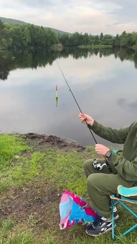 A person wearing a green tracksuit and a hat is sitting on a blue folding chair next to a calm lake, holding a fishing rod. A colorful plastic bag is on the ground in front of them. The person is fishing, with the rod extended over the water. The lake reflects the surrounding trees and the cloudy sky. The ground around the lake is covered in grass and some mud. A yellow and red fishing float is visible in the water.