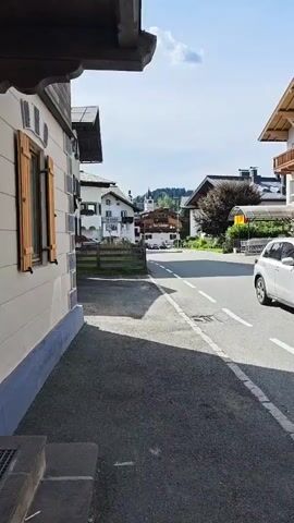 The video shows a street scene in a European village or town. The foreground is a paved sidewalk next to a street. On the right side of the street, there is a multi-story building with wooden balconies and large windows. Several cars are parked in front of the building. The street itself has white dashed lines indicating lanes. In the background, there are other buildings, some with traditional architecture, and trees. A church steeple is visible in the distance, along with rolling hills or mountains. The sky is blue with a few scattered clouds, and the sun is casting shadows. The overall impression is a sunny day in a picturesque, possibly alpine, setting.