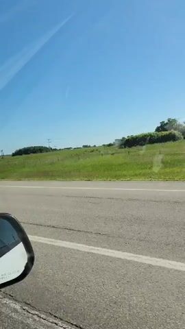 The video shows a view from inside a car, looking out the passenger window. The car is on a paved road with a white line on the right side. The landscape outside is a rolling green hill under a clear blue sky. There are a few utility poles and some trees scattered across the hill. The side mirror of the car is visible in the lower left corner of the frame. The video appears to be filmed while the car is in motion.