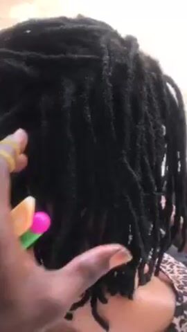 Here's a description of the video:
The video shows a person with black dreadlocks. A hand is seen using a comb with colorful accents to style the dreadlocks. The person being styled is wearing a leopard print top. The background appears to be a light-colored surface, possibly a blanket or sheet. The camera angle focuses closely on the hair styling process.