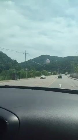 The video is a first-person view from inside a vehicle driving on a road. The road has multiple lanes and is lined with traffic cones and poles. On the left side of the road, there are trees and a mountain in the background with a building on it. On the right side of the road, there are more trees and a building. Several cars are visible on the road. The sky is cloudy with some blue visible. The video is short and appears to be a continuous shot.