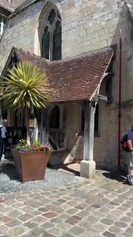 The video shows the exterior of an old stone building with a wooden porch and a tiled roof. A tall, spiky green plant is in a large brown planter in front of the porch. A cobblestone path leads up to the building. A person in a striped shirt and dark pants stands to the left of the planter, facing away from the camera. Another person, wearing a backpack and a blue shirt, stands to the right of the porch, facing the building. The sky is blue and clear.