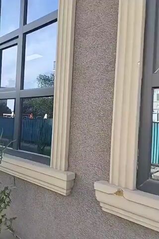A video of a window and the wall of a building. The window has a grey frame and is divided into multiple panes by black mullions. The glass reflects a building, a fence, and trees. The wall of the building is textured and has a decorative column next to the window.