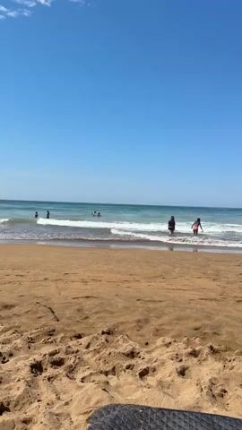 The video shows a beach scene under a clear blue sky. In the foreground, a sandy beach with some footprints is visible. In the middle ground, waves are crashing on the shore, and several people are in the water. Two individuals, one wearing black and the other wearing pink, are standing in the shallow water, facing the sea. In the background, more people are swimming or playing in the ocean. The water is a greenish-blue color. A dark object, possibly a towel or mat, is partially visible in the bottom right corner of the frame.