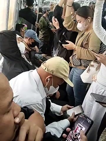 The video shows the interior of a crowded train car. Numerous passengers are standing and holding onto overhead bars. Most are wearing face masks. Several individuals are looking at their mobile phones. One man in the foreground, wearing a light-colored shirt and a tan baseball cap, is looking down at his phone. To his left, another person, partially visible, is also looking at a phone. In the background, passengers are packed closely together. The lighting is artificial and appears somewhat dim.