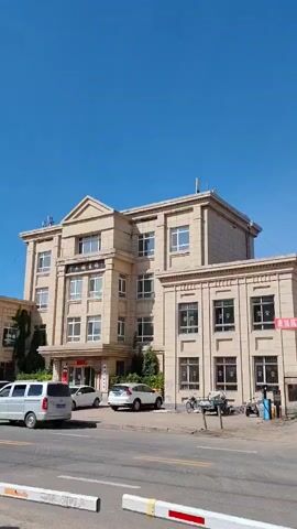 A multi-story tan building stands against a clear blue sky. The building has many windows and an arched entranceway. Several cars are parked in front of the building, and a white van is on the left side of the frame. A red and white barrier is visible in the foreground. The video zooms in slightly on the building.