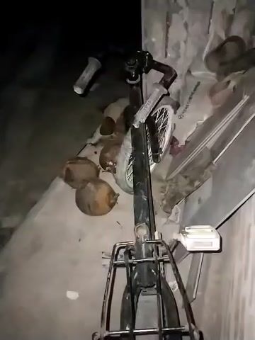 The video shows a black bicycle indoors. The lighting is poor, making the scene dimly lit. Next to the bicycle, there are about four brown, round objects, possibly coconuts. The background also includes some items covered by a white cloth. The video does not show any people or significant actions. The camera angle is low, looking up at the bicycle, and the scene appears static.